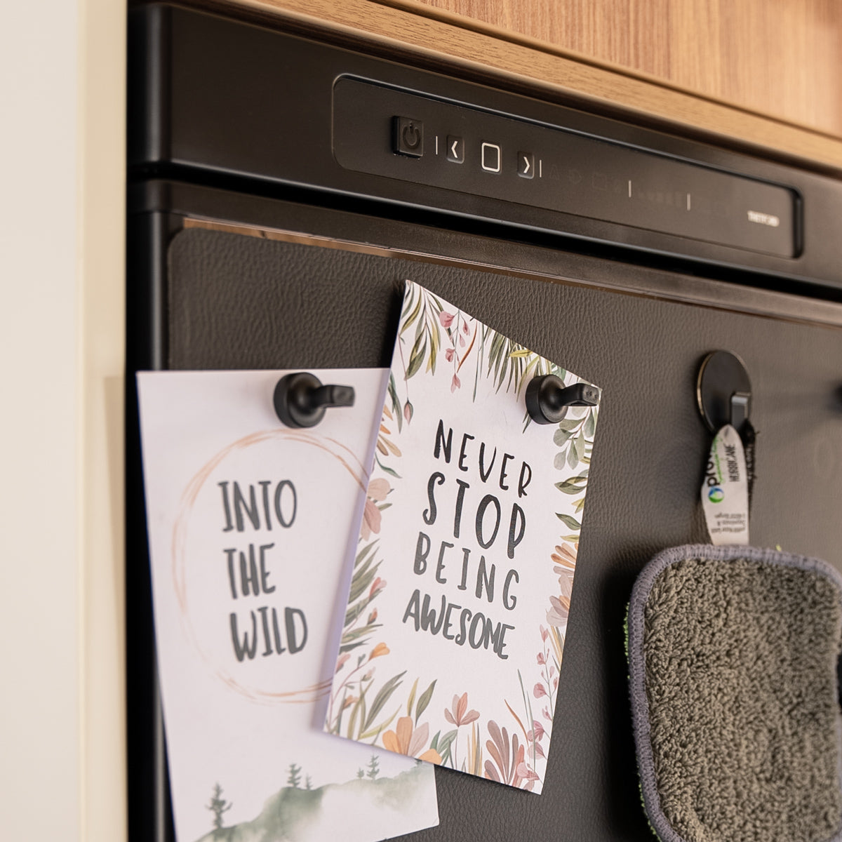 Karten "Into the wild" und "Never stop being awesome" sowie Topflappen mit Magnet-Pins und Magnet-Haken an Wand angebracht